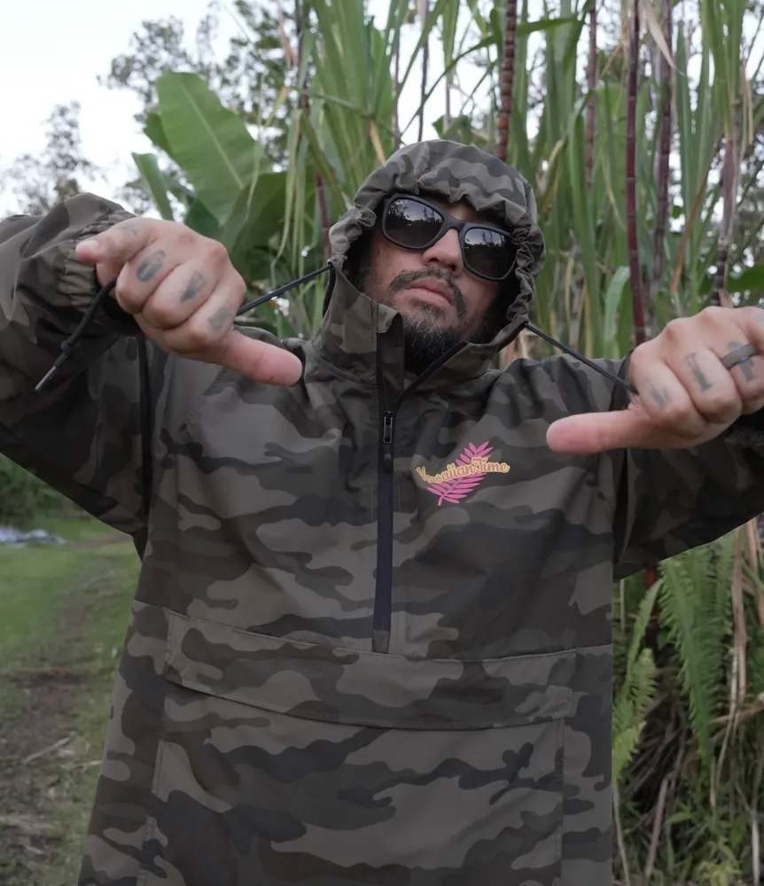 A man in a camo hooded jacket and dark sunglasses points both thumbs at the Hawaiian Time logo on his chest, with sugarcane and ferns behind him.