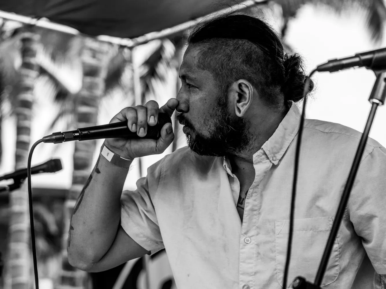 A black and white photo of a man in a button-up shirt singing close into a microphone, with palm trees behind him.