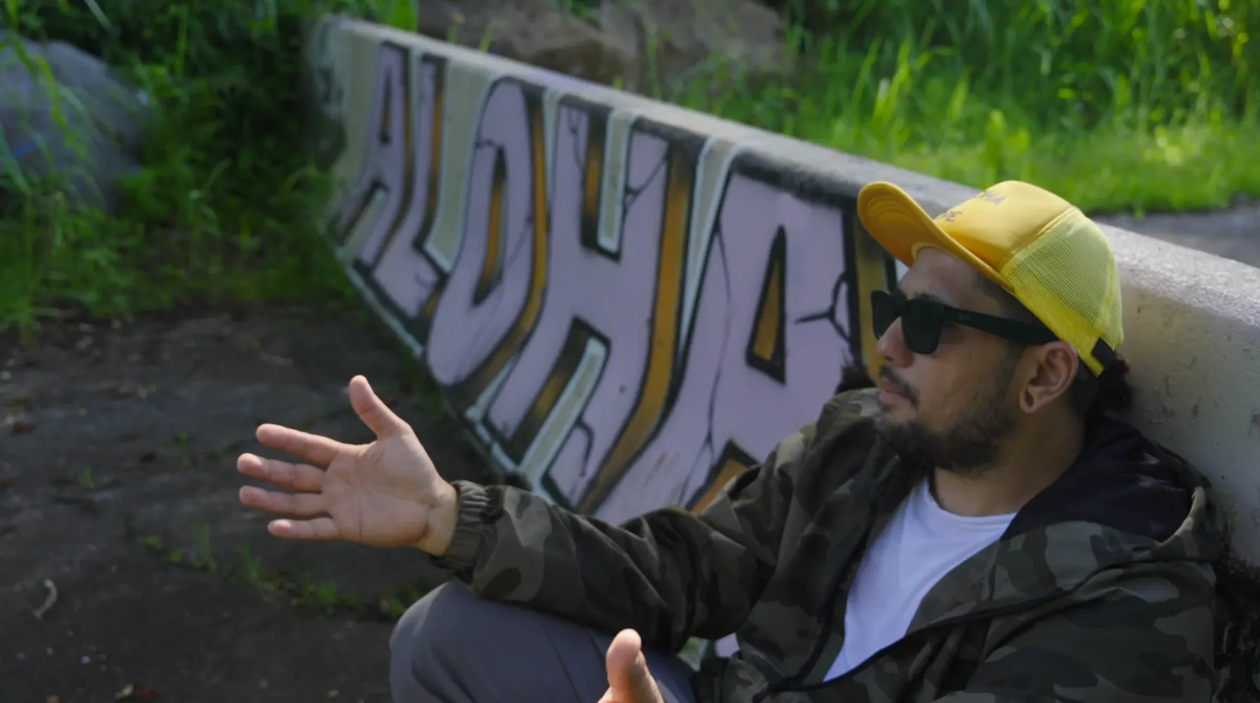 A man in a yellow cap and sunglasses sits against a low wall painted with the word Aloha, one hand open as he talks.