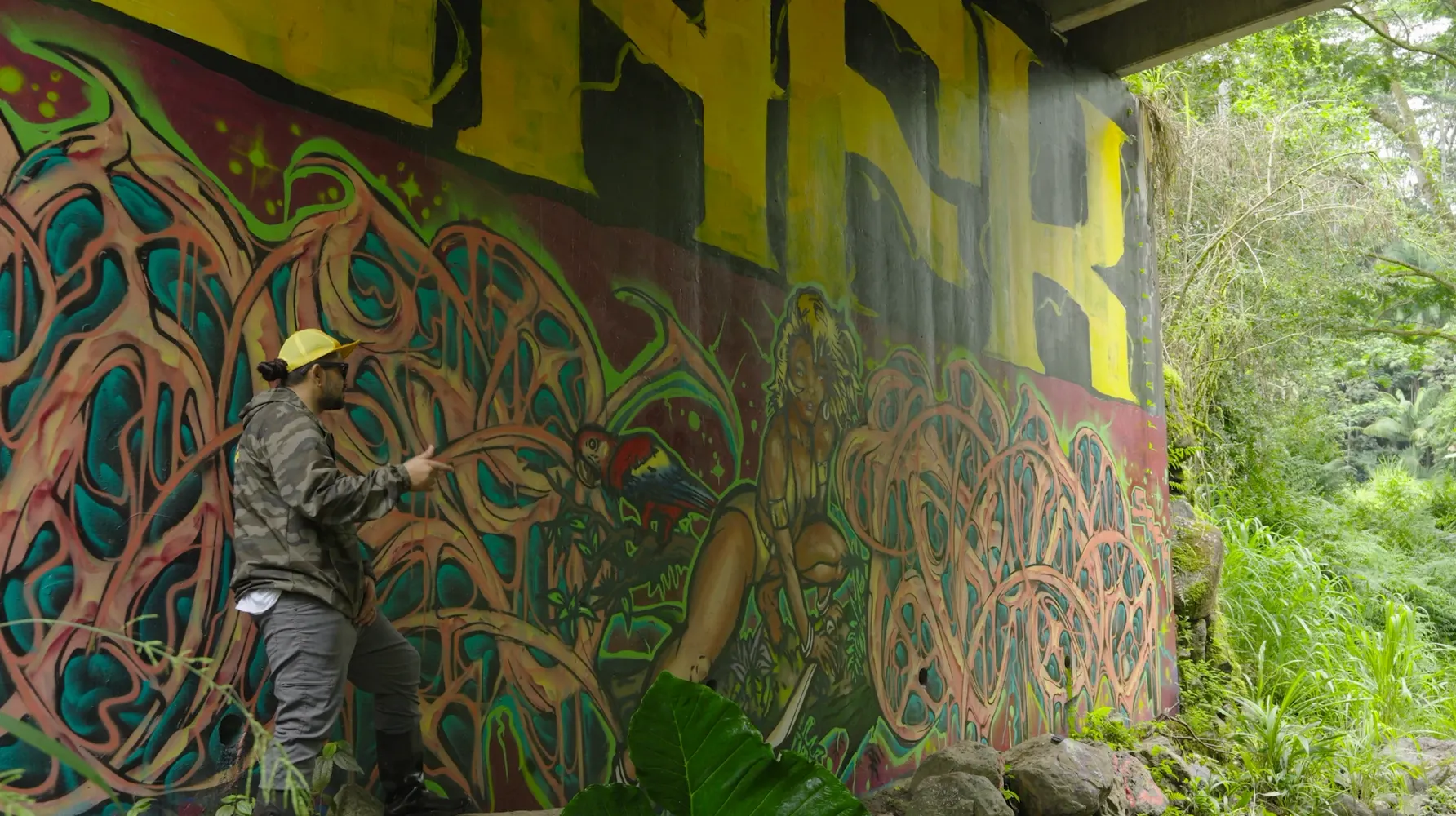A man in a yellow cap and camo jacket points toward a large colorful mural painted under a bridge, with green forest beside it.